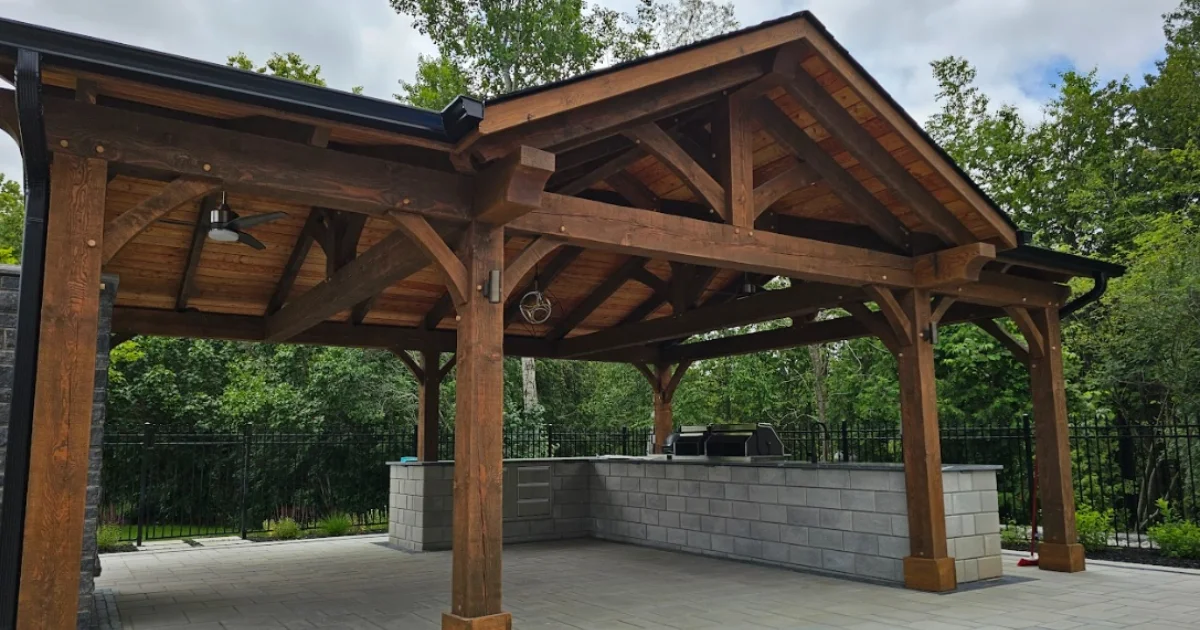 Covered timber pavilion featuring an outdoor kitchen with built-in grill and prep counter, surrounded by trees.